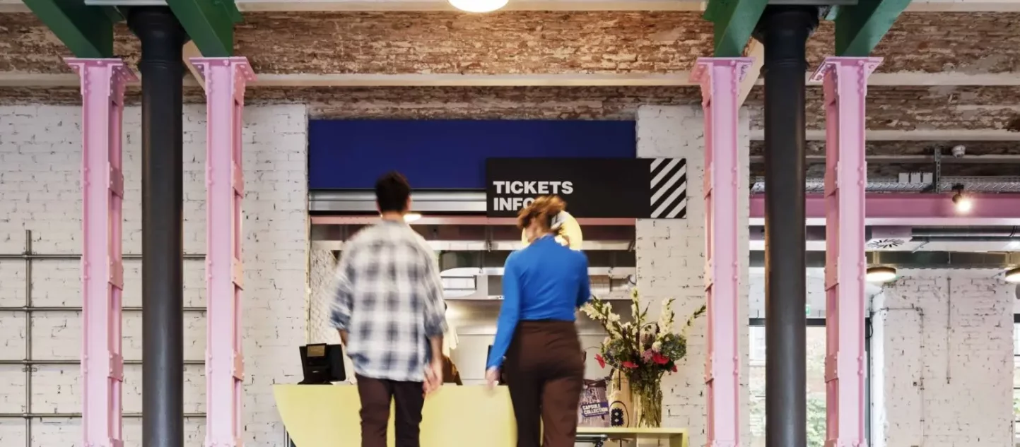 Zwei Personen laufen auf ein Schild mit der Aufschrift 'Tickets, Info' zu in einem Raum mit bunten Stahltr&auml;gern und unverputzten Decken